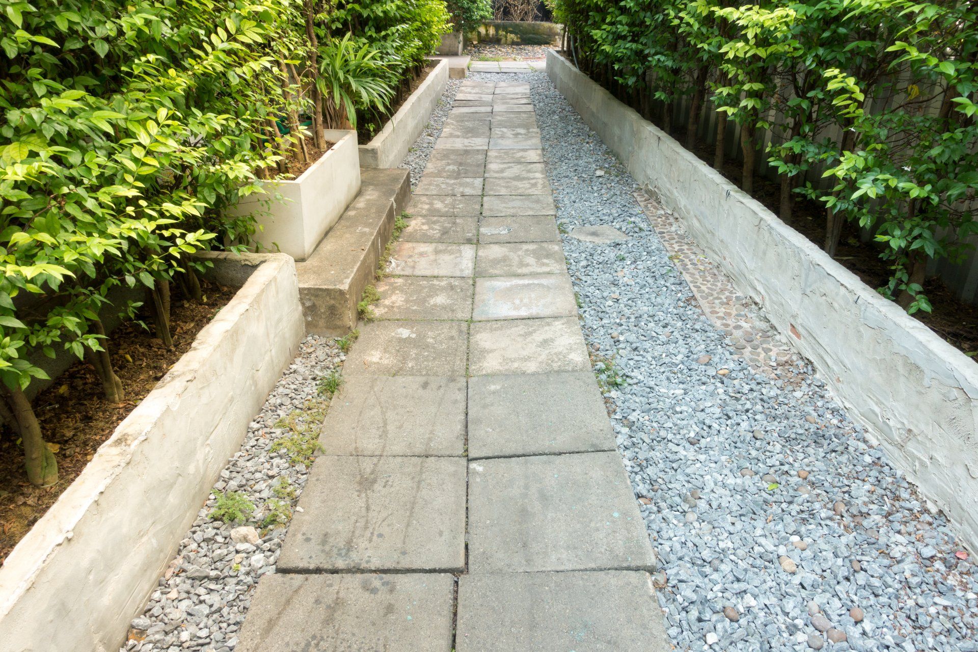Stone walkway bordered by lush green bushes, with gravel on one side and concrete on the other